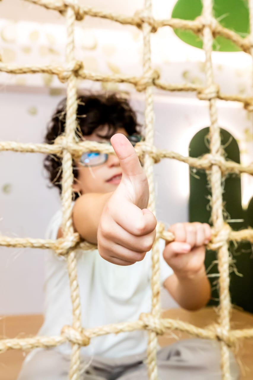 Criança fazendo sinal de positivo através da rede de escalada